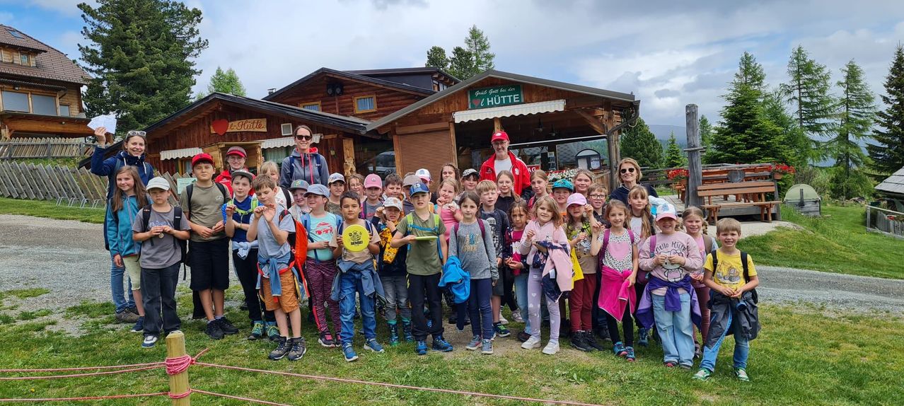 Eine Gruppe von Kindern und Erwachsenen steht vor einem Holzgebäude. Das Gebäude hat ein Schild mit der Aufschrift 'Griss Gac bei der Hutte.' Einige Kinder halten Frisbees.