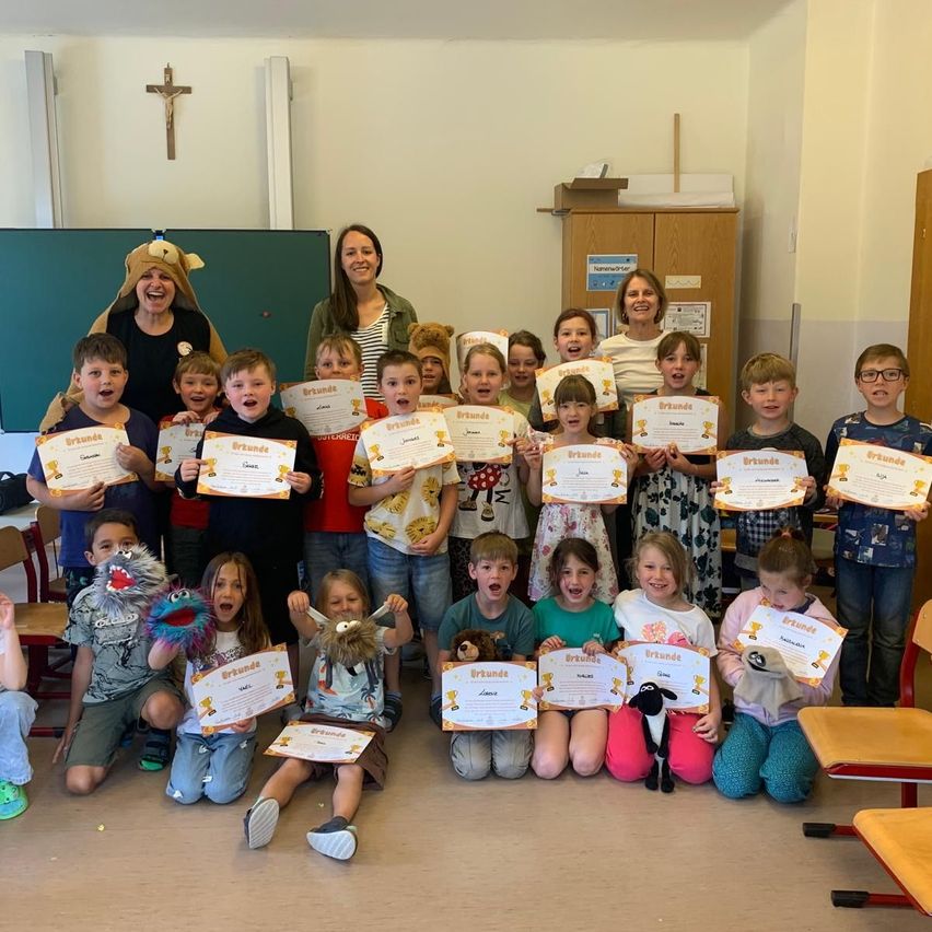 Eine Gruppe von Kindern und Erwachsenen posiert mit Zertifikaten in einem Klassenzimmer. Einige knien, während andere hinter ihnen stehen.