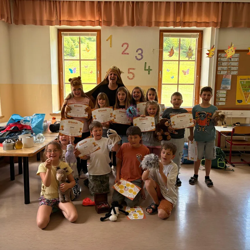 Klassenfoto von Kindern mit Urkunden, einer Lehrerin in Löwenkostüm und festlicher Klassenzimmer-Atmosphäre mit Dekorationen und Zahlen an der Wand.