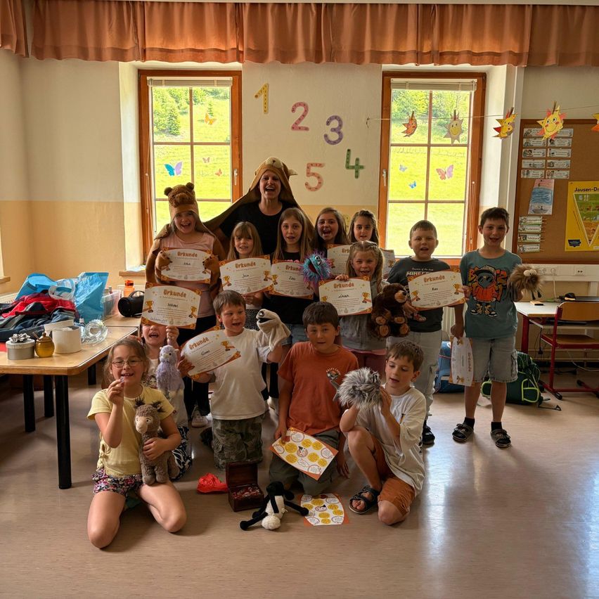 Klassenfoto von Kindern mit Urkunden, einer Lehrerin in Löwenkostüm und festlicher Klassenzimmer-Atmosphäre mit Dekorationen und Zahlen an der Wand.