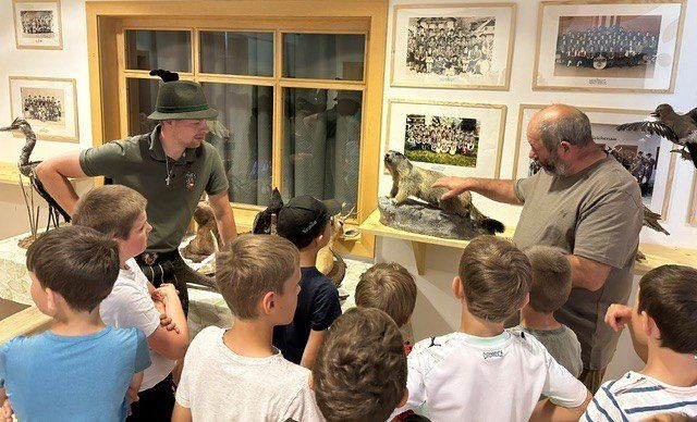 Zwei Männer stehen vor einer Gruppe von Kindern, erklären und zeigen ausgestopfte Tiere auf einem Tisch. Der Raum hat gerahmte Bilder an den Wänden.