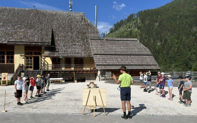 Ein Mann spricht zu einer Gruppe von Kindern vor einem rustikalen Gebäude mit einem Berg im Hintergrund.