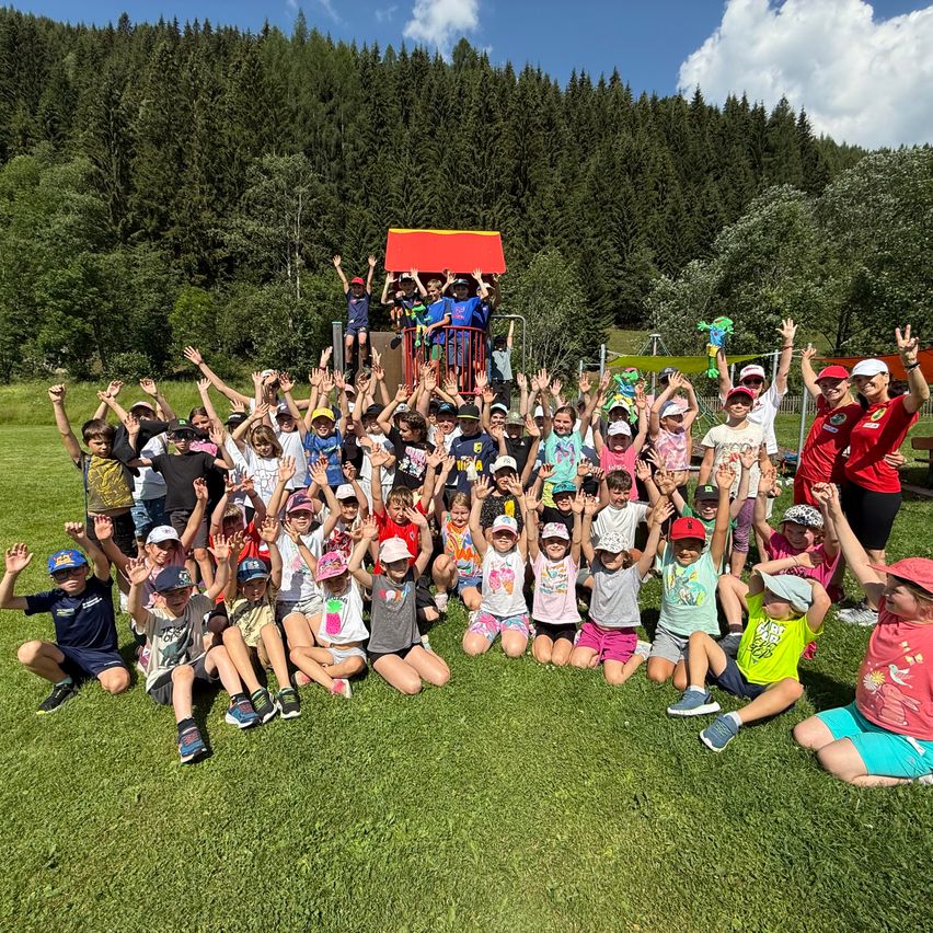 Eine Gruppe von Kindern posiert für ein Foto auf einem Rasen mit Bäumen und einem Spielplatz im Hintergrund. Einige Kinder stehen und andere sitzen. Alle tragen Hüte und Turnschuhe. Eine Frau in einem roten Shirt steht hinter ihnen.