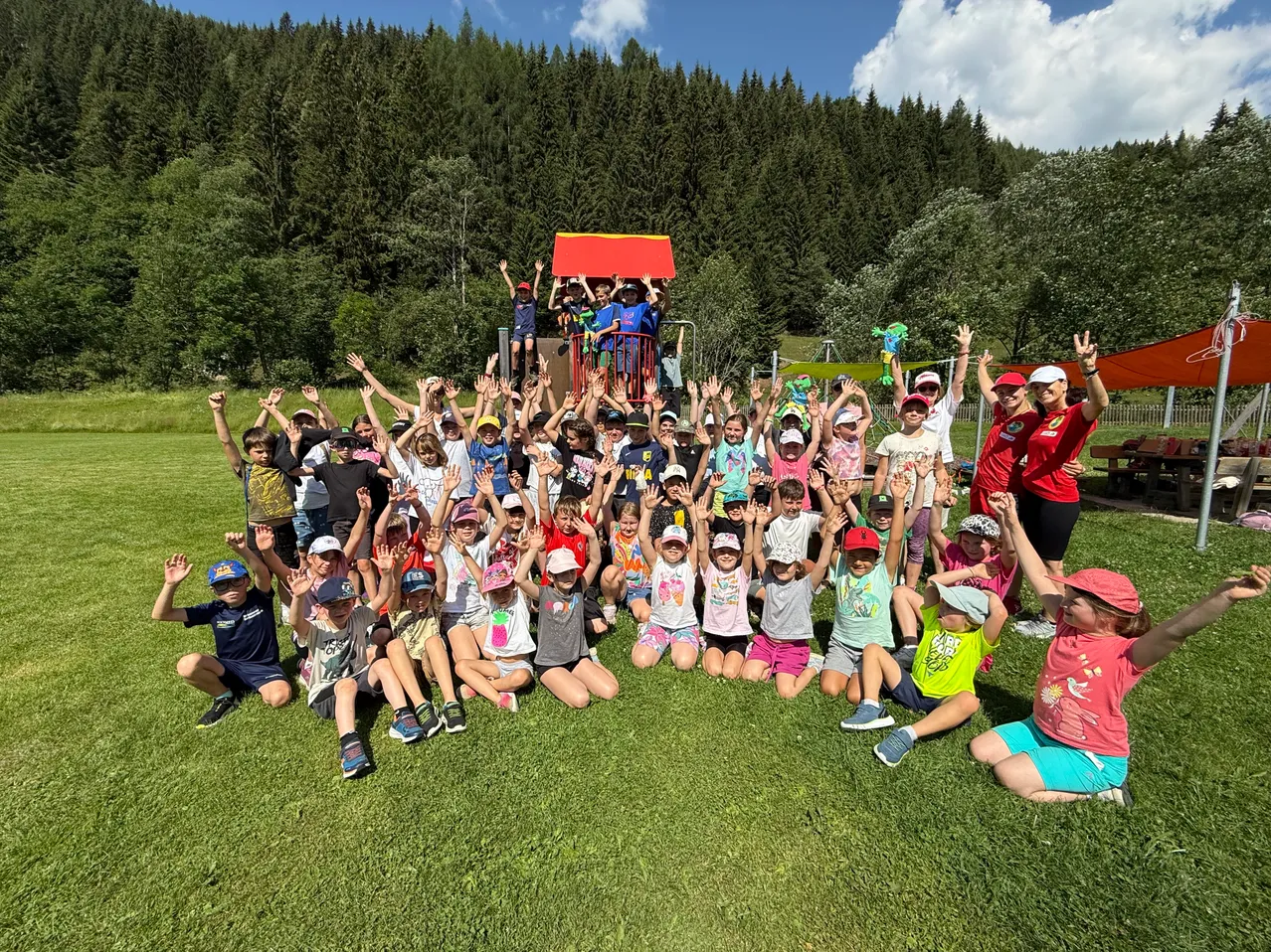 Eine Gruppe von Kindern posiert für ein Foto auf einem Rasen mit Bäumen und einem Spielplatz im Hintergrund. Einige Kinder stehen und andere sitzen. Alle tragen Hüte und Turnschuhe. Eine Frau in einem roten Shirt steht hinter ihnen.