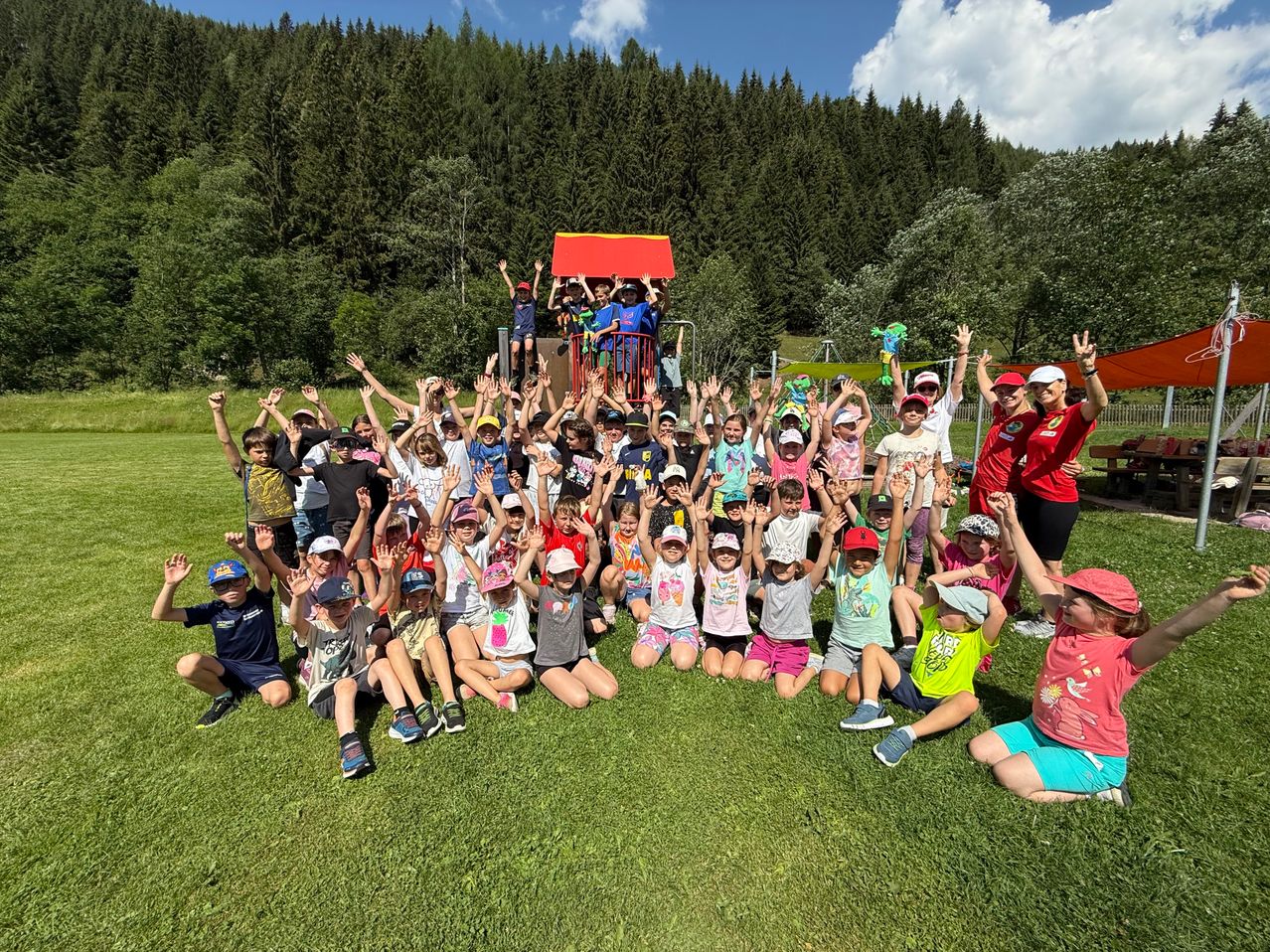 Eine Gruppe von Kindern posiert für ein Foto auf einem Rasen mit Bäumen und einem Spielplatz im Hintergrund. Einige Kinder stehen und andere sitzen. Alle tragen Hüte und Turnschuhe. Eine Frau in einem roten Shirt steht hinter ihnen.