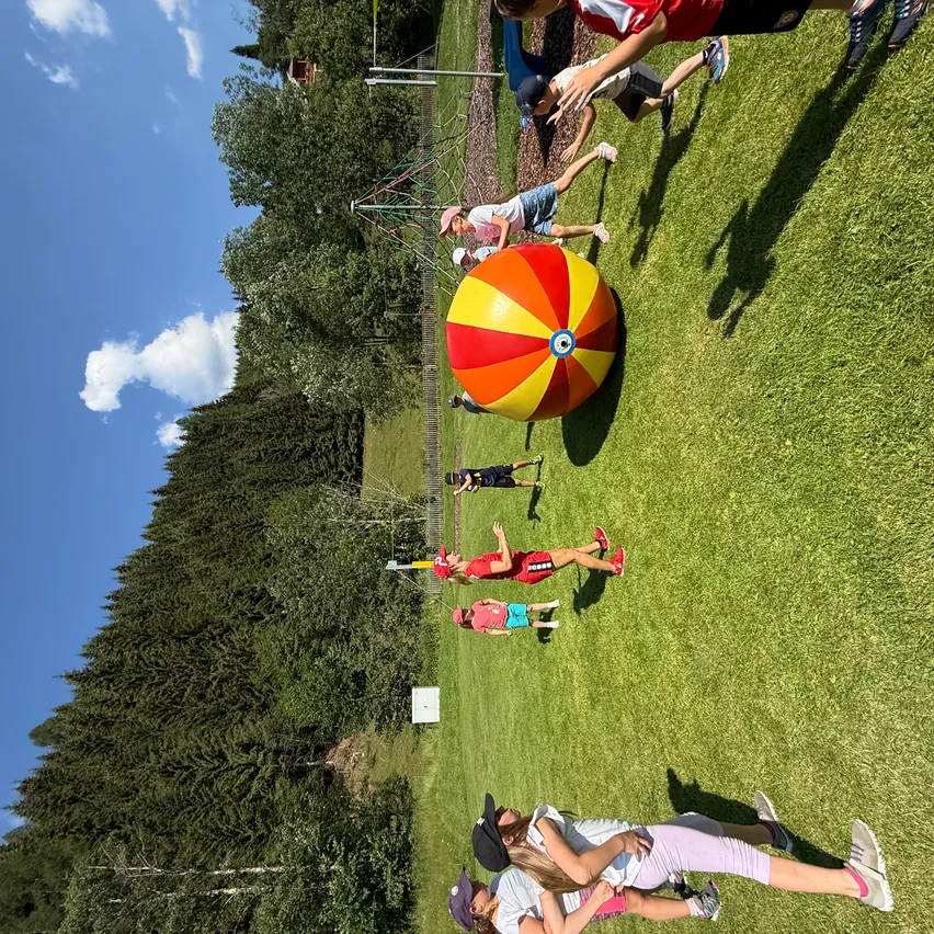 Eine Gruppe von Kindern spielt mit einem großen mehrfarbigen Ball auf einem grünen Feld unter einem blauen Himmel.