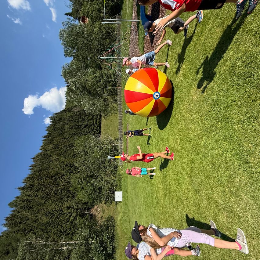 Eine Gruppe von Kindern spielt mit einem großen mehrfarbigen Ball auf einem grünen Feld unter einem blauen Himmel.