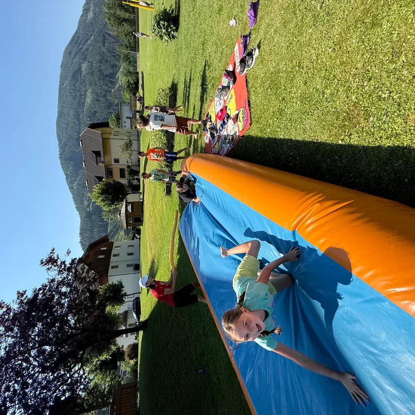 Ein Mädchen rutscht auf einer farbenfrohen aufblasbaren Rutsche auf einem Rasenfeld, mit Gebäuden und Bergen im Hintergrund.