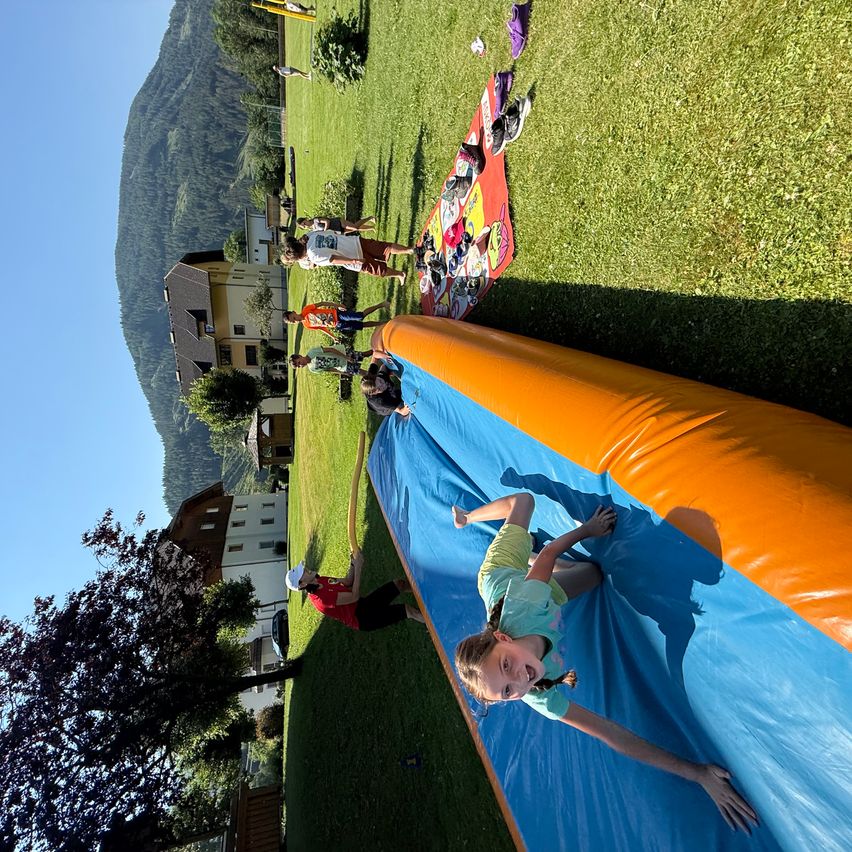 Ein Mädchen rutscht auf einer farbenfrohen aufblasbaren Rutsche auf einem Rasenfeld, mit Gebäuden und Bergen im Hintergrund.