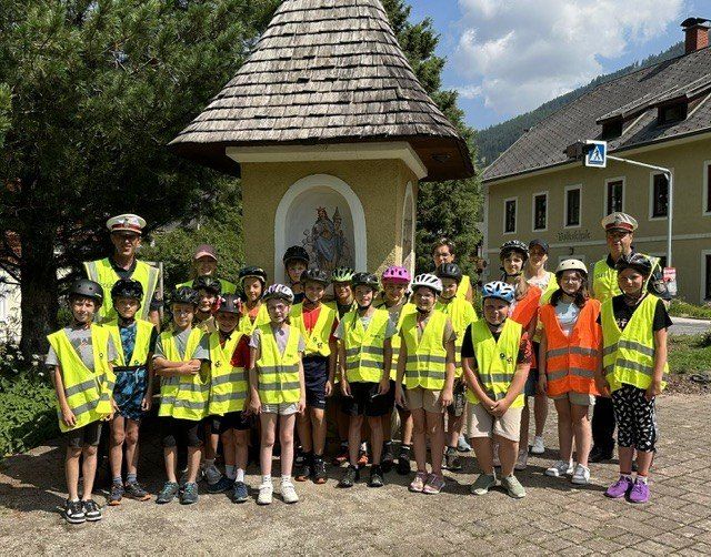 Eine Gruppe von Kindern in leuchtenden Westen und Helmen steht draußen und posiert für ein Foto vor einer kleinen Kapelle.