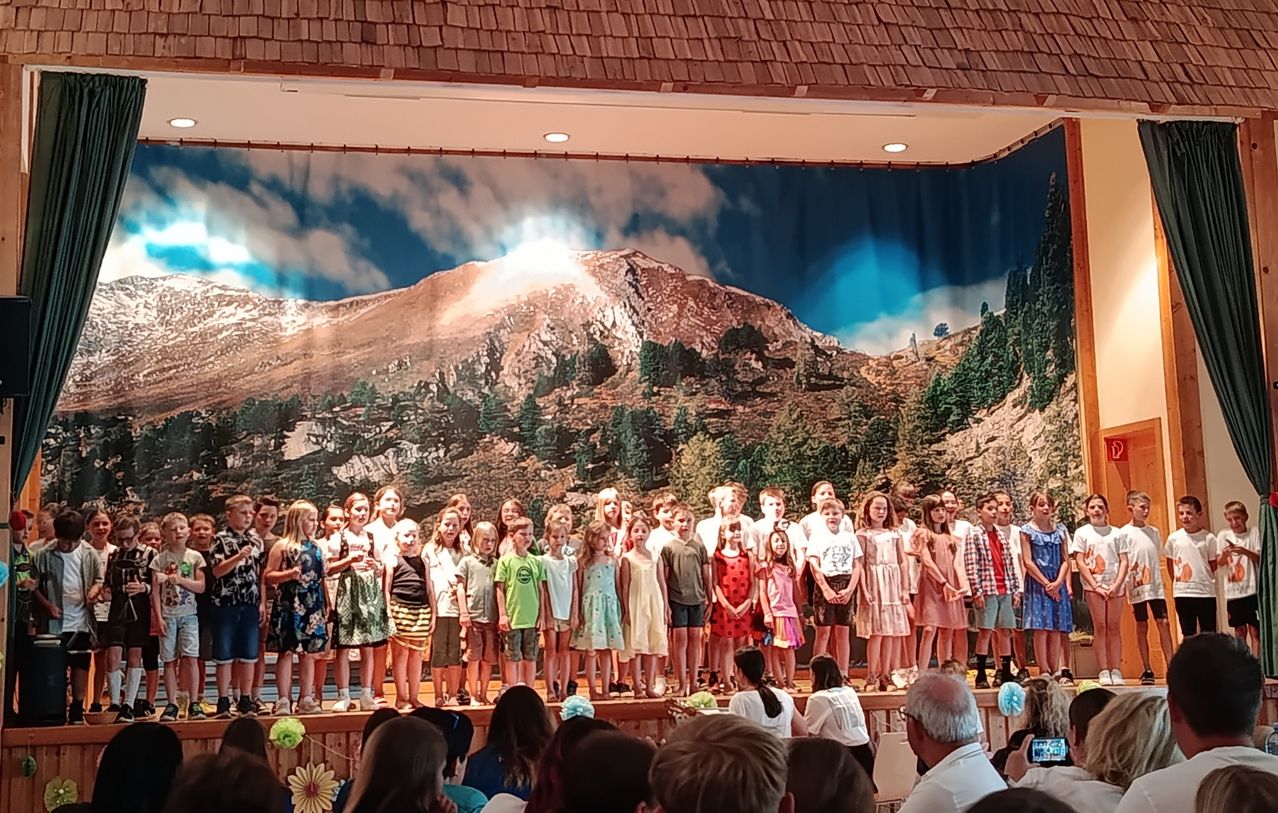 Eine Gruppe von Kindern in Sommerkleidung steht auf einer Bühne mit einem Hintergrund von Bergen und Bäumen. Sie treten wahrscheinlich für ein Publikum auf.