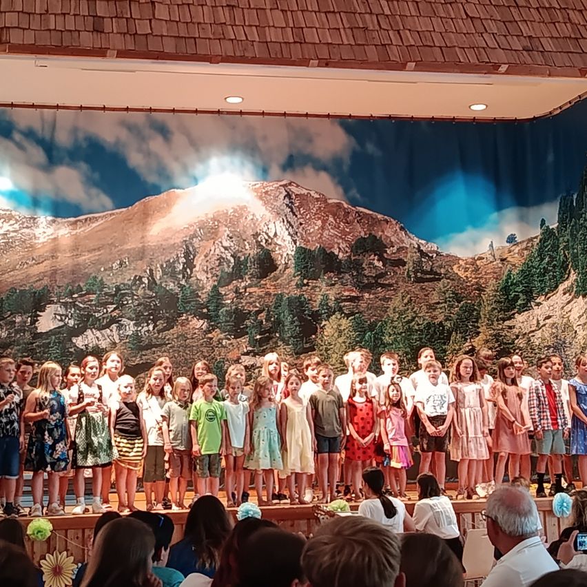 Eine Gruppe von Kindern in Sommerkleidung steht auf einer Bühne mit einem Hintergrund von Bergen und Bäumen. Sie treten wahrscheinlich für ein Publikum auf.