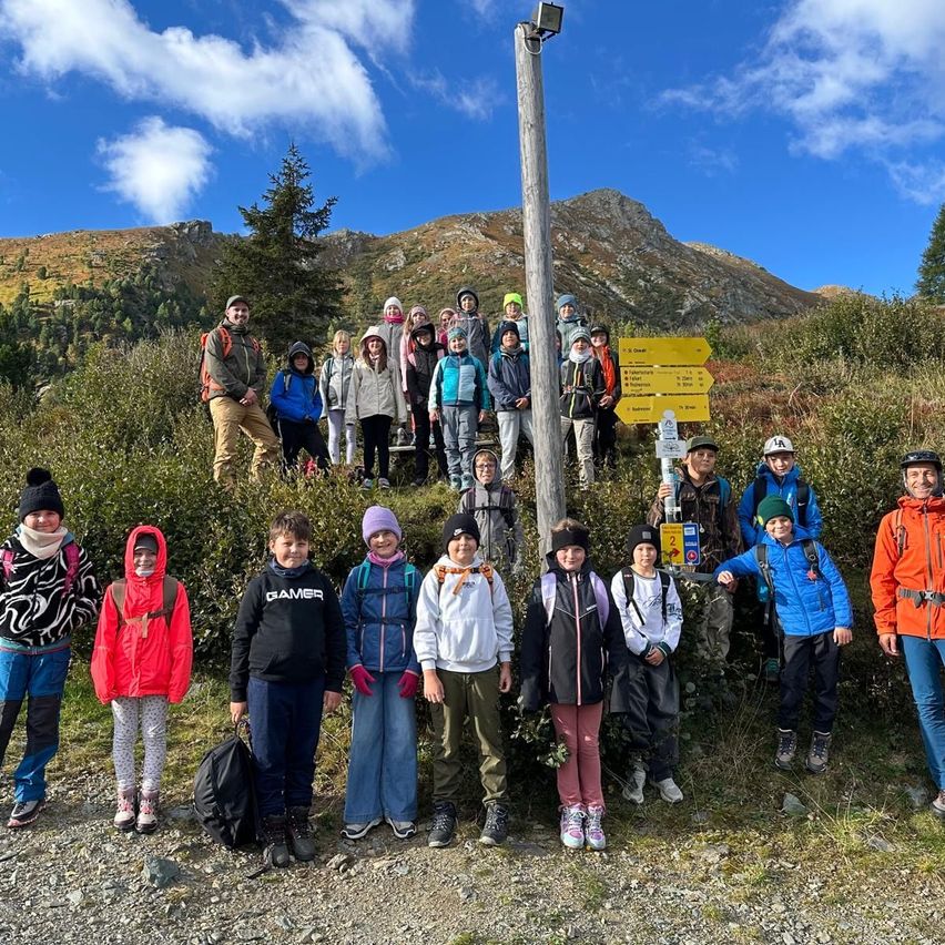 Eine Gruppe von Wanderern, einige mit Rucksäcken, posiert für ein Foto auf einem Bergpfad mit einem Schild dahinter.