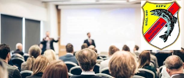 Eine Konferenz mit Teilnehmern in Reihen. Zwei Redner stehen auf der Bühne. Ein Wappen mit Text befindet sich auf der rechten Seite des Bildes.