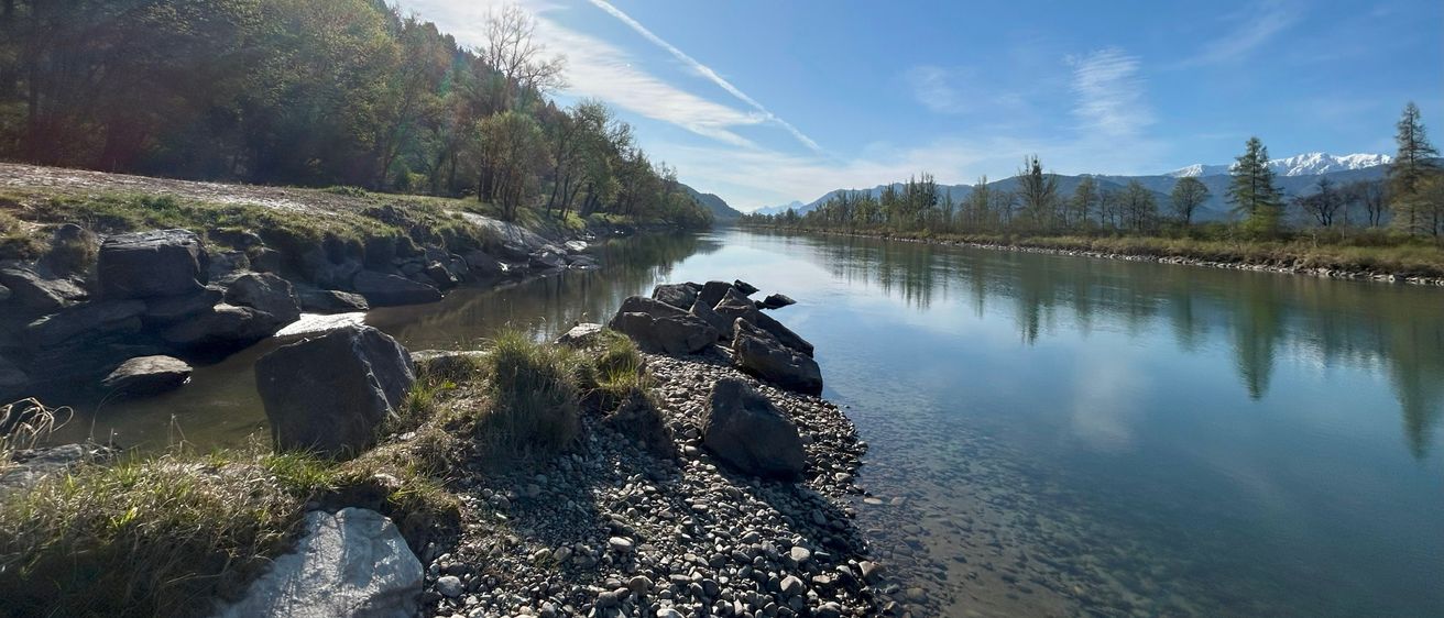 Ein Fluss unter einem strahlenden Sonnenschein, flankiert von felsigen Ufern, mit Bäumen und Bergen in der Ferne.