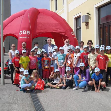 Eine Gruppe von Kindern, Erwachsenen und zwei Männern posiert für ein Foto vor einem roten Zelt mit OBB-Logo. Sie tragen Hüte und Turnschuhe, und einige haben Taschen dabei. Das Gebäude dahinter hat Glasfenster und eine Überwachungskamera.