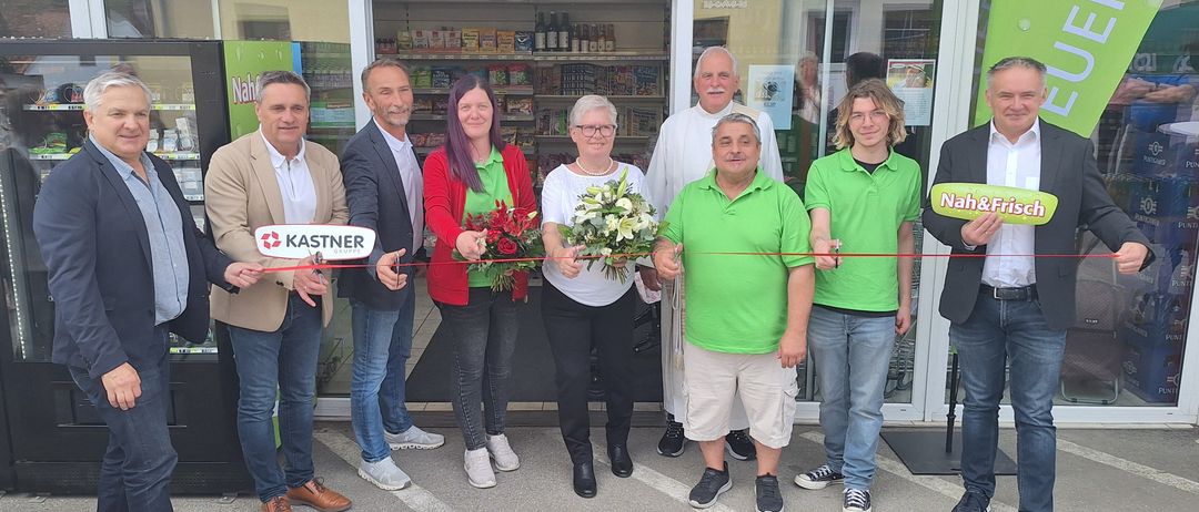 Menschen posieren vor einem Geschäft, das sie mit einem Band halten. Ein Mann hält einen Blumenstrauß. Das Geschäft hat eine Glastür.
