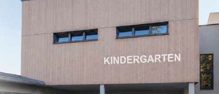 Die Vorderseite eines Kindergartengebäudes mit dem Wort Kindergarten über dem Eingang. Es gibt drei Topfpflanzen und zwei Fenster am Gebäude.