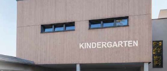 Die Vorderseite eines Kindergartengebäudes mit dem Wort Kindergarten über dem Eingang. Es gibt drei Topfpflanzen und zwei Fenster am Gebäude.