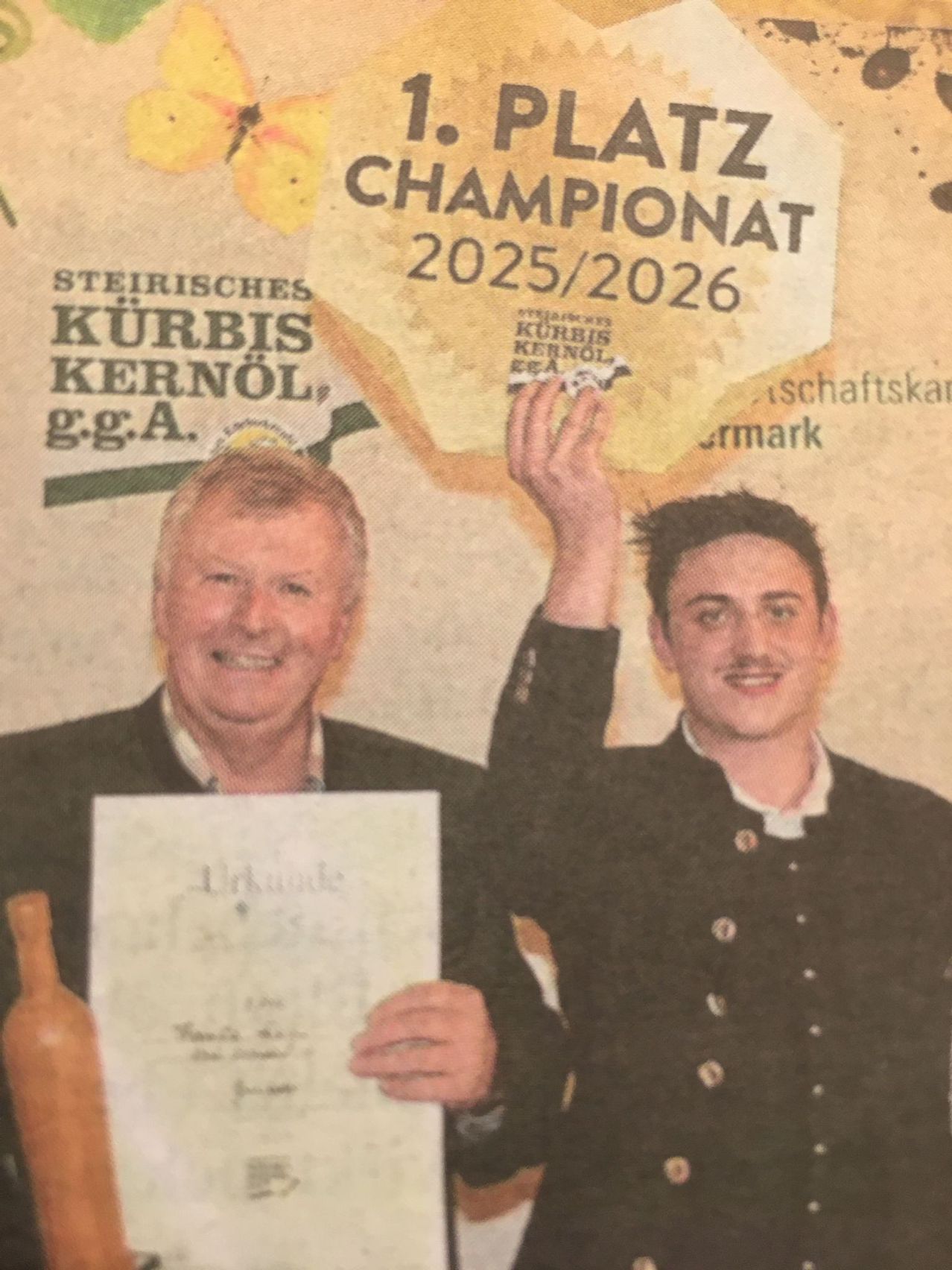 Zwei Männer in formeller Kleidung stehen lächelnd und halten ein Zertifikat und einen Kürbis. Der Mann auf der linken Seite hält ein Zertifikat mit der Aufschrift 'Urkunde'. Hinter ihnen befindet sich ein Plakat mit deutschem Text.