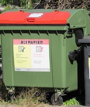 Ein grüner Abfallbehälter mit rotem Deckel und einem Aufkleber mit der Aufschrift 'Altpapier', wahrscheinlich für die Papierrecycling. Er steht auf Rädern und ist im Freien aufgestellt.