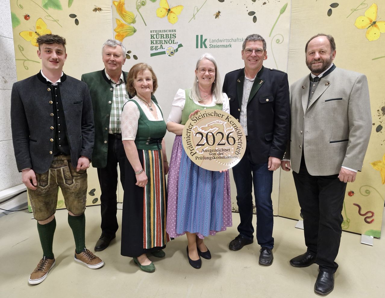 A group of people stand in front of a green banner. A woman holds a plaque with the text 2026. The man on the right wears a suit, and the man on the left wears a traditional outfit.