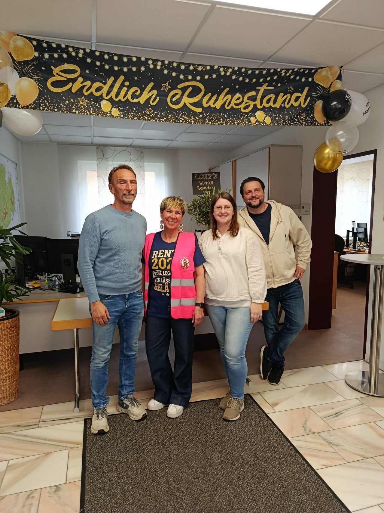 Four people stand in an office, smiling. A woman in the middle wears a reflective vest and a shirt saying 'REN 2020'. A banner above reads 'Endlich Ruhestand'. Behind them is a desk with a monitor.