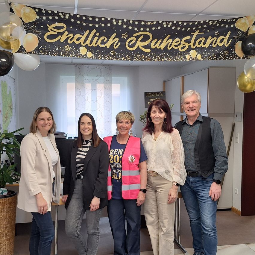 Five people are standing in front of a wall with a golden and black banner that says 'Endlich Ruhestand'. They are smiling for a photo. Behind them are balloons and a potted plant.