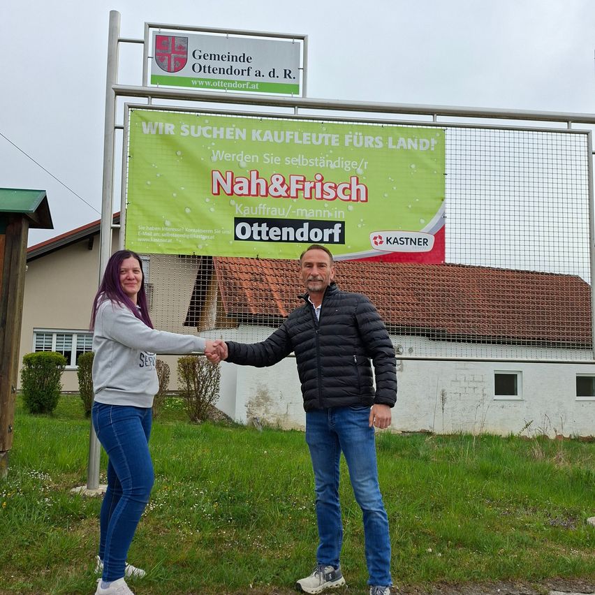 A man and a woman shake hands in front of a banner advertising a local market. The banner reads Nah & Frisch Ottendorf. Behind them, a building with a red roof and a fence is visible.