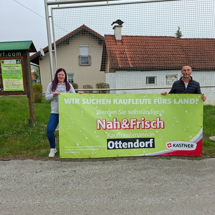 Two people stand behind a banner advertising local produce. The woman wears purple hair and the man has a beard. A house is in the background.