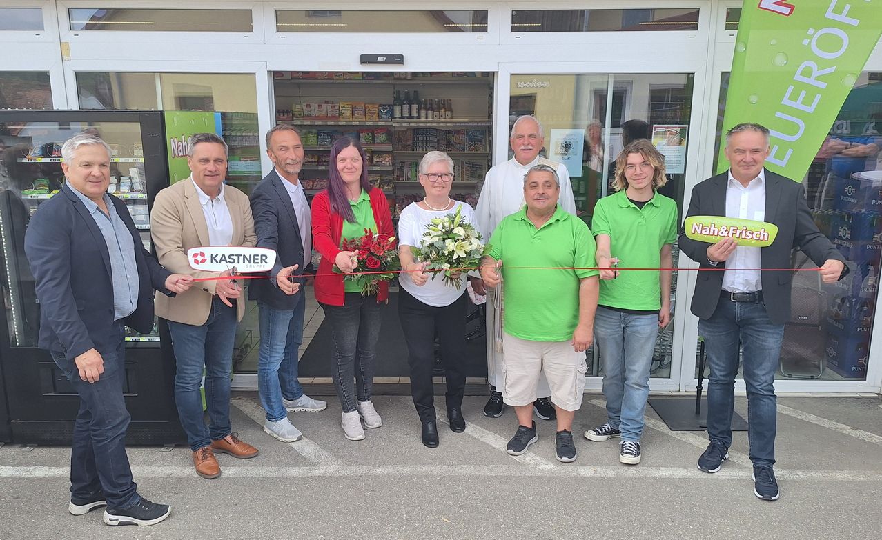 A group of people in green and white shirts holding flowers stand in front of a store, cutting a ribbon.