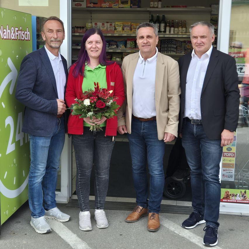 Four people stand in front of a store, smiling and posing for a photo. A woman in the center holds a bouquet of flowers.