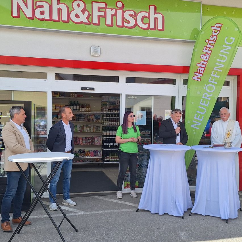 Five people stand in front of a store with 'Nah&Frisch' on the sign. Two are speaking into microphones at tables with white cloths. Inside, shelves full of products are visible.