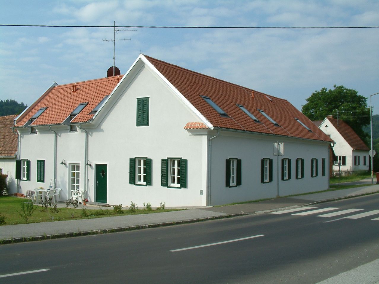 A white house with a brown roof and green shutters. It is situated on a street with a curb.