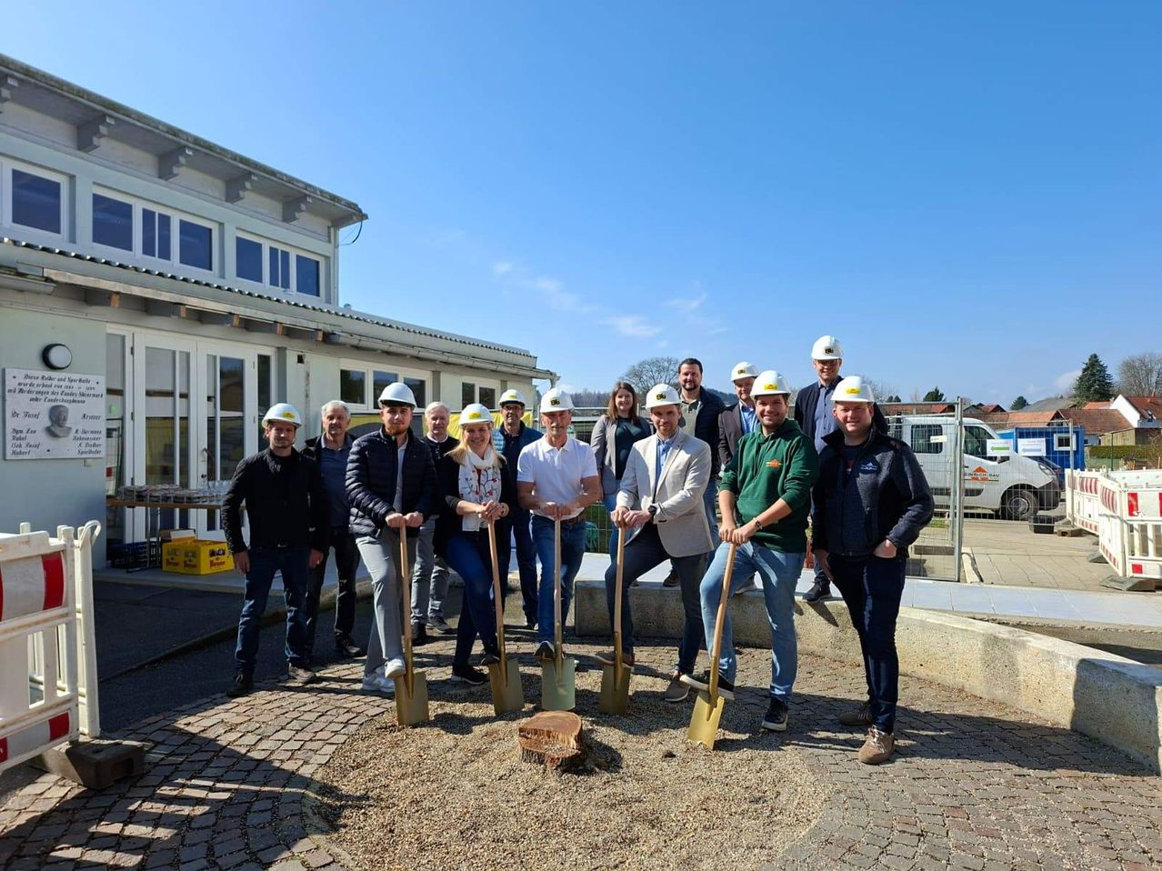 Eine Gruppe von Menschen in Schutzhelmen posiert mit Schaufeln für ein Foto vor einem Gebäude.