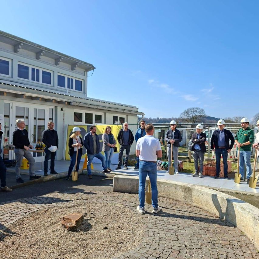Eine Gruppe von Menschen in Helmen steht um eine Baustelle herum. Sie halten Schaufeln und diskutieren wahrscheinlich ein Projekt. Das Gebäude hinter ihnen hat viele Fenster und eine gelbe Fahne.