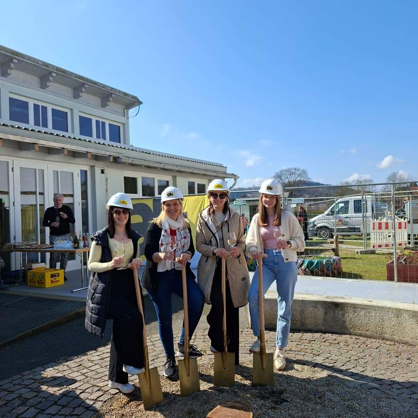 Fünf Frauen in Schutzhelmen halten Schaufeln und posieren für ein Foto vor einem Gebäude. Hinter ihnen steht ein Mann am Tisch mit Flaschen, und ein Mann telefoniert. Im Hintergrund befinden sich ein Zaun und geparkte Fahrzeuge.
