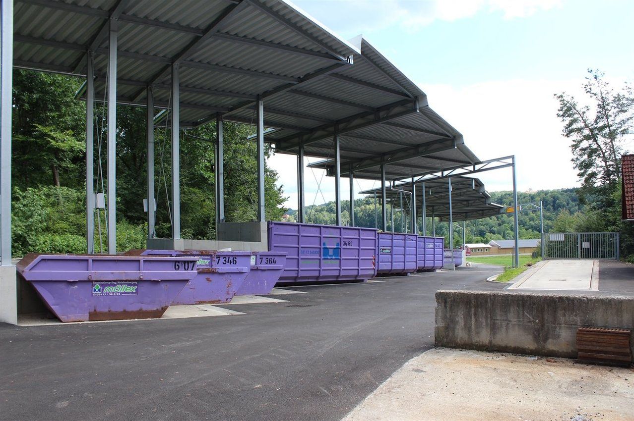 A row of purple dumpsters under metal roofs, numbered 607, 7346, and 7364, on a paved road with trees and mountains in the background.