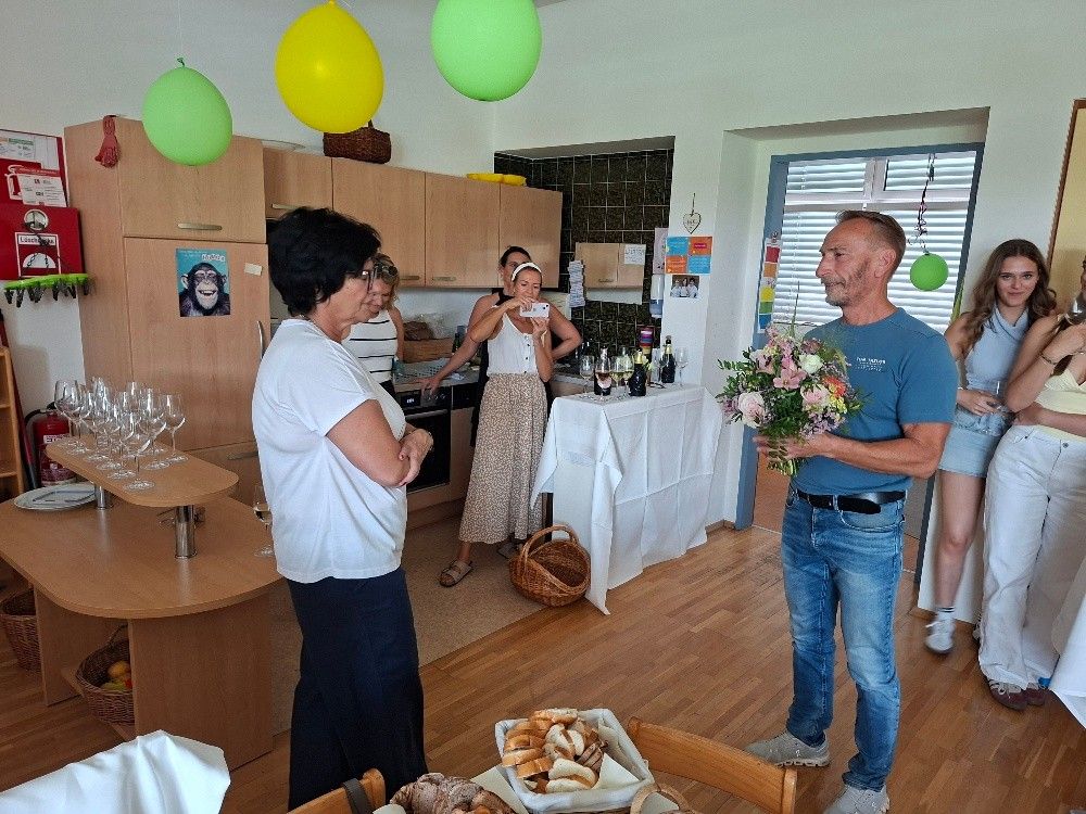 A group of people in a kitchen, a woman holding a bouquet of flowers, and a man holding a camera. A table with bread and wine bottles is in the foreground.