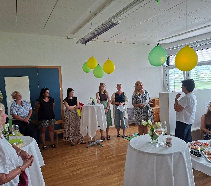 A group of women stand around tables in a room with balloons hanging from the ceiling. Some hold papers, and one woman speaks to the group.