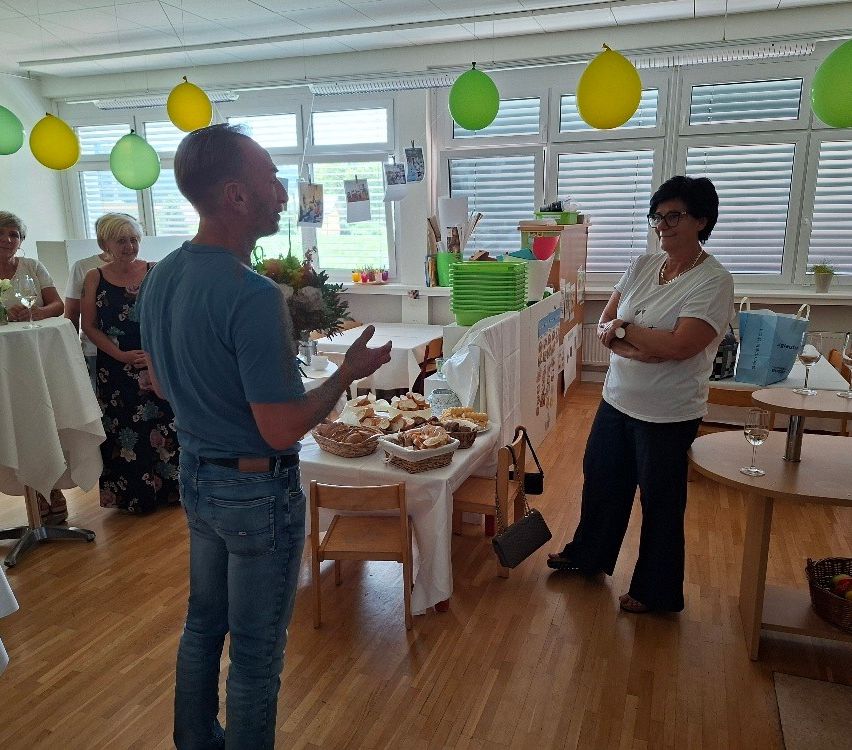 A group of people are standing in a room with a table full of food, balloons, and a woman wearing glasses and a watch.