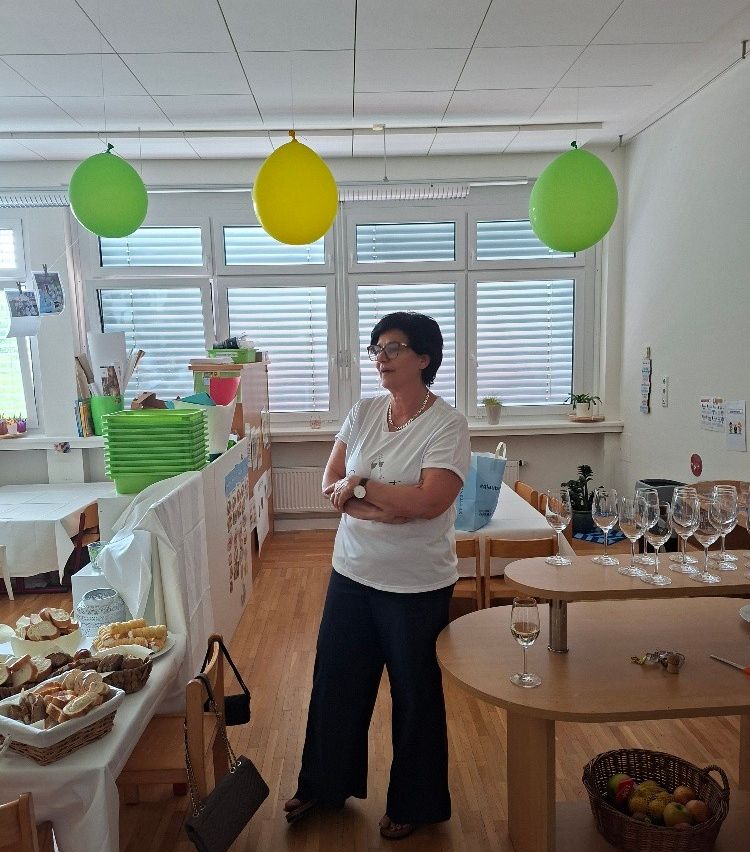 A woman stands in a room with balloons, a table with bread and wine glasses, and a window with blinds.