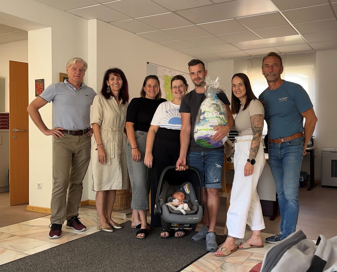 Eine Gruppe von Menschen steht in einem Raum, einer hält ein Baby in einem Kinderwagen und ein anderer ein Geschenkkörbchen.