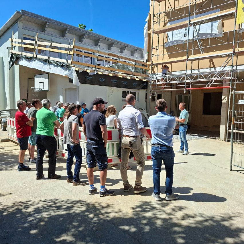 Eine Gruppe von Menschen steht vor einem im Bau befindlichen Gebäude. Sie stehen hinter Barrieren, und eine Person ist auf einem Gerüst. Es sind weitere Personen in der Umgebung, einige unterhalten sich.