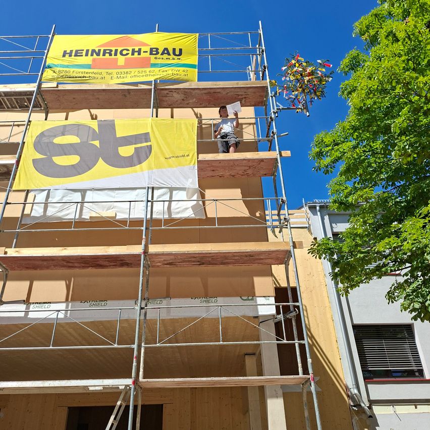 Eine Person steht auf einem Gerüst an der Seite eines Gebäudes mit Bannern, die 'Heinrich-Bau' und 'st' lesen. Der Himmel ist klar.