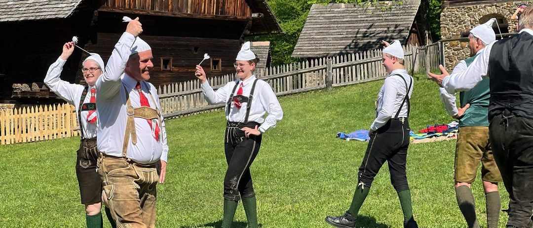 Eine Gruppe von Männern in traditioneller bayerischer Kleidung führt auf einer Rasenfläche vor alten Holzhäusern auf. Sie halten weiße Pfeile in den Händen und scheinen an einer Aktivität beteiligt zu sein.