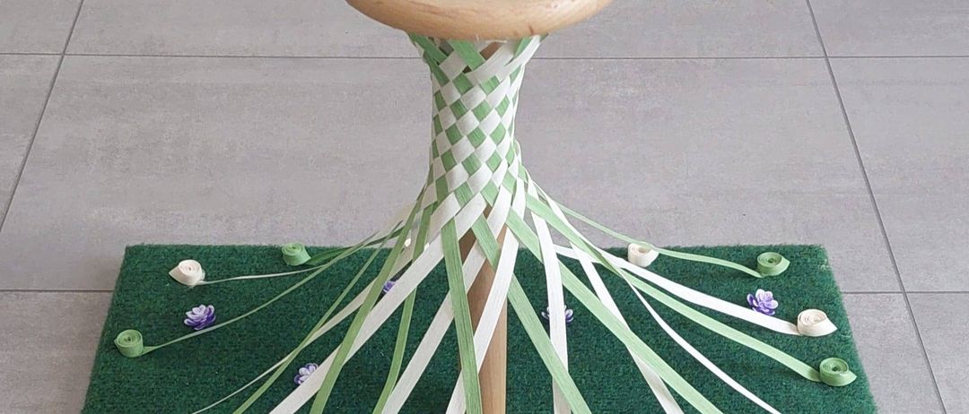 Ein dekorativer hölzerner Stuhl mit Bändern und Blumen steht auf einem grünen Teppich, der ein Wappen mit 'Geimatmercin' und '01.MAI 2025' zeigt. Die Basis ist kunstvoll mit Bändern und Blumen gewoben.