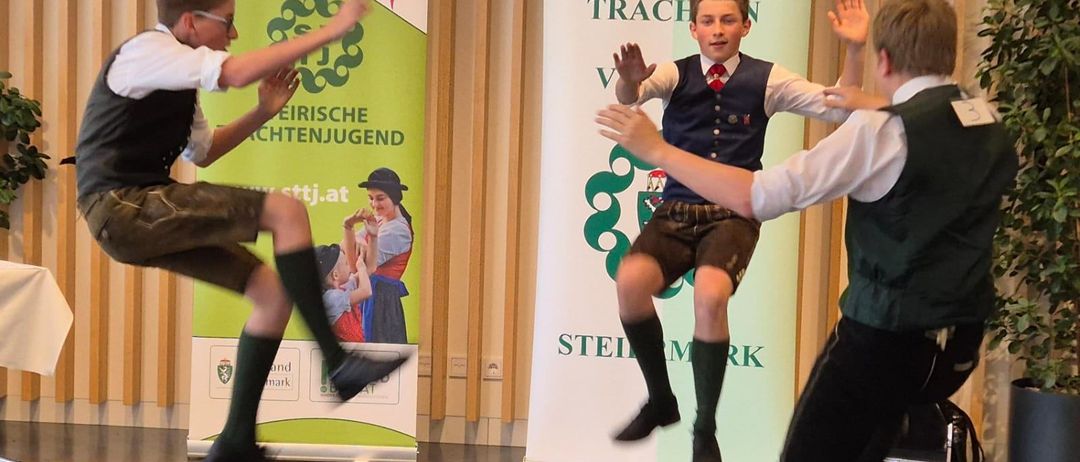 Vier Jungen in traditionellen Kostümen führen einen Tanz vor einem Schild mit der Aufschrift 'Landes Trachen' auf. Sie springen und heben ihre Hände. Dahinter stehen zwei Banner, eines mit grünem Hintergrund und das andere weiß.