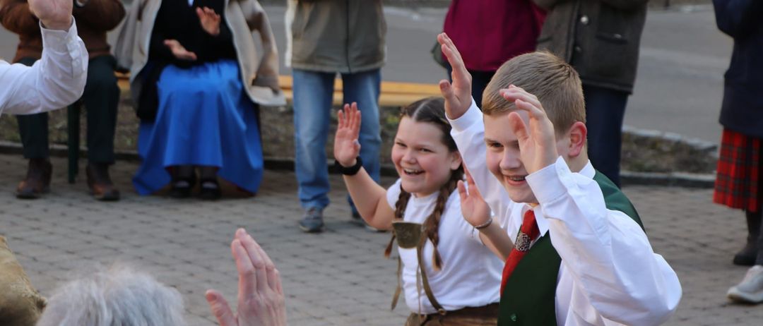 Eine Gruppe von Menschen, darunter Kinder in traditioneller Kleidung, klatschen und lächeln im Freien.