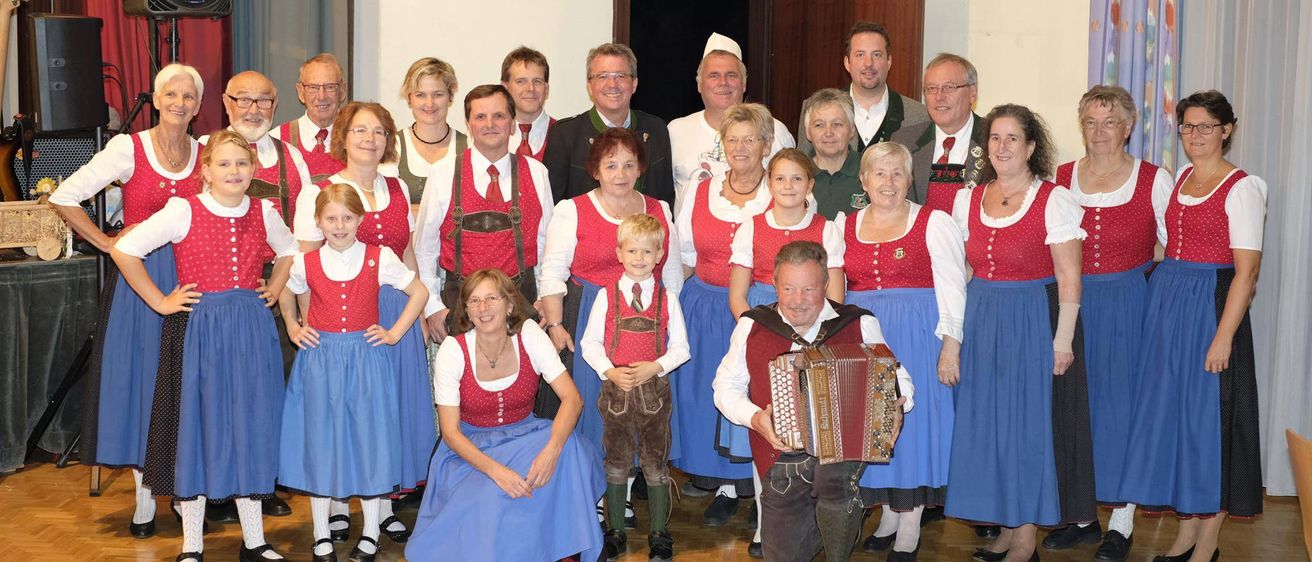 Eine Gruppe von Menschen in traditioneller bayerischer Kleidung posiert für ein Foto in einem Raum. Ein Mann spielt Akkordeon, während andere im Hintergrund stehen und sitzen.